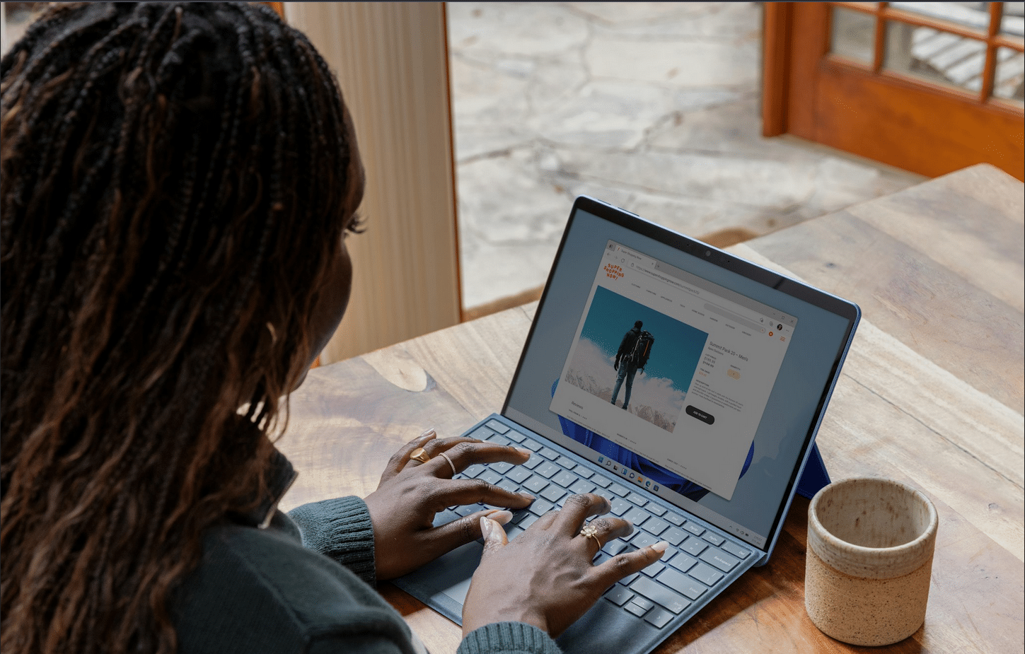 A person with braided hair uses a laptop on a wooden table, browsing an online store featuring a man in a coat, perhaps for their next tabletop RPG session. A ceramic mug sits to the right, while a window and door create a cozy, immersive backdrop.